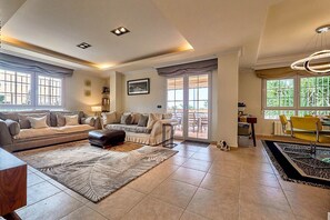 A spacious living room with a sofa set, a coffee table, and a rug. There is a dining area with a table and chairs, and a view of the outdoors through large windows.