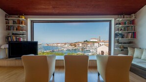 Una habitación con un amplio ventanal que da a un puerto deportivo y edificios, un televisor y estanterías llenas de libros.