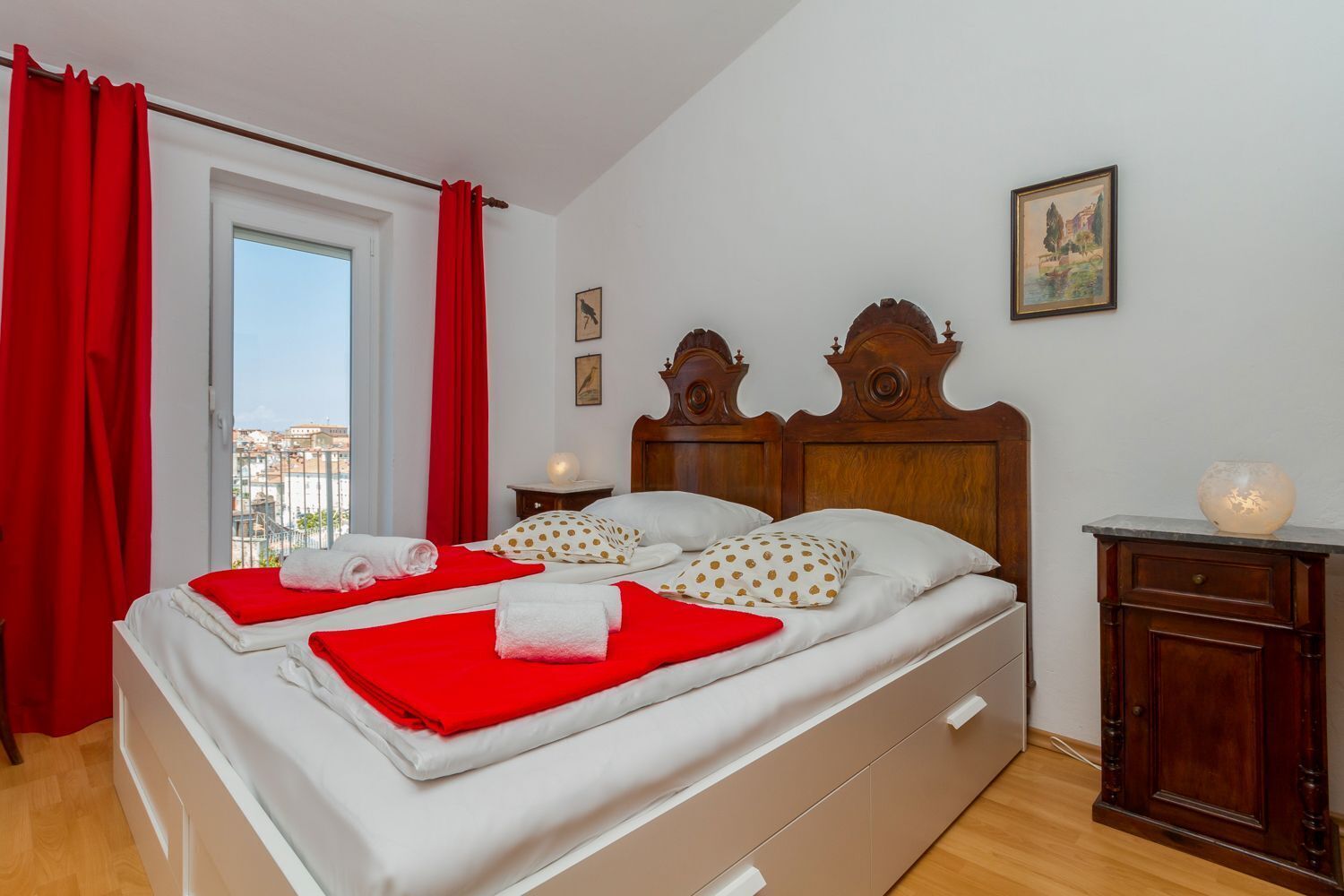 Ein Doppelbett mit roter und weißer Bettwäsche, ein Holz-Kopfstück und Blick auf Gebäude durch ein Fenster mit roten Vorhängen.