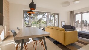 A modern living room with a dining table, sofa, and chairs. There is a balcony with a railing and a view of trees.
