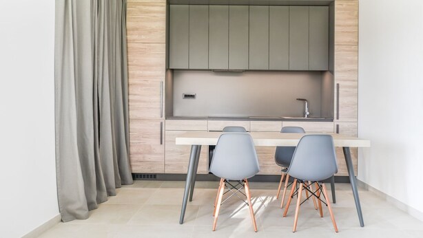 A compact kitchen with a built-in stove, a sink, and a small countertop. There are two grey chairs and a white table.
