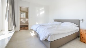 A bedroom with a bed, a wooden nightstand, a window with curtains, and a bathroom with a mirror and shelves.
