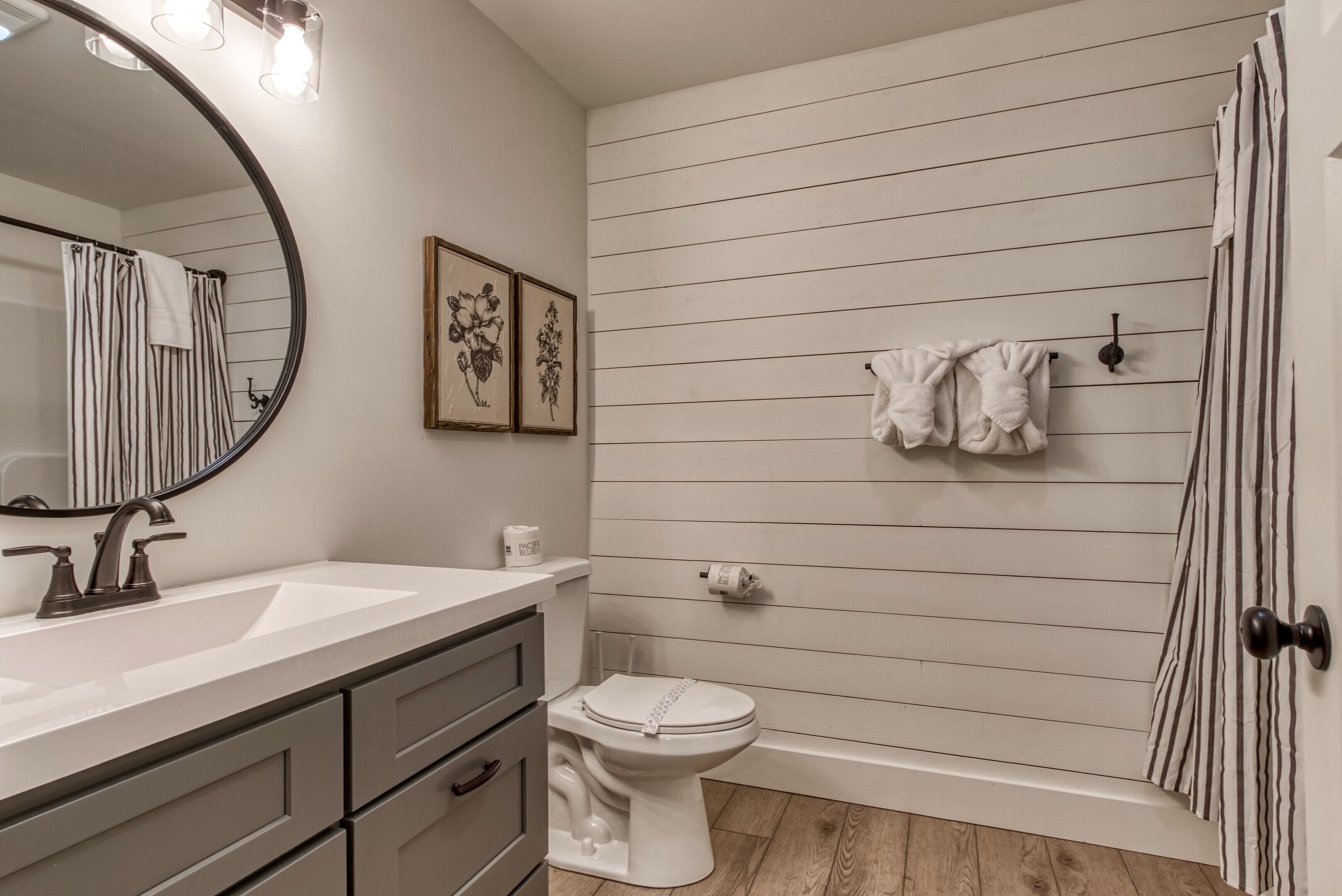 Een badkamer met een wit wastafel, toilet en douche met gestreepte gordijnen. Er hangen afbeeldingen in lijsten aan de muur en een handdoekrek met handdoeken.