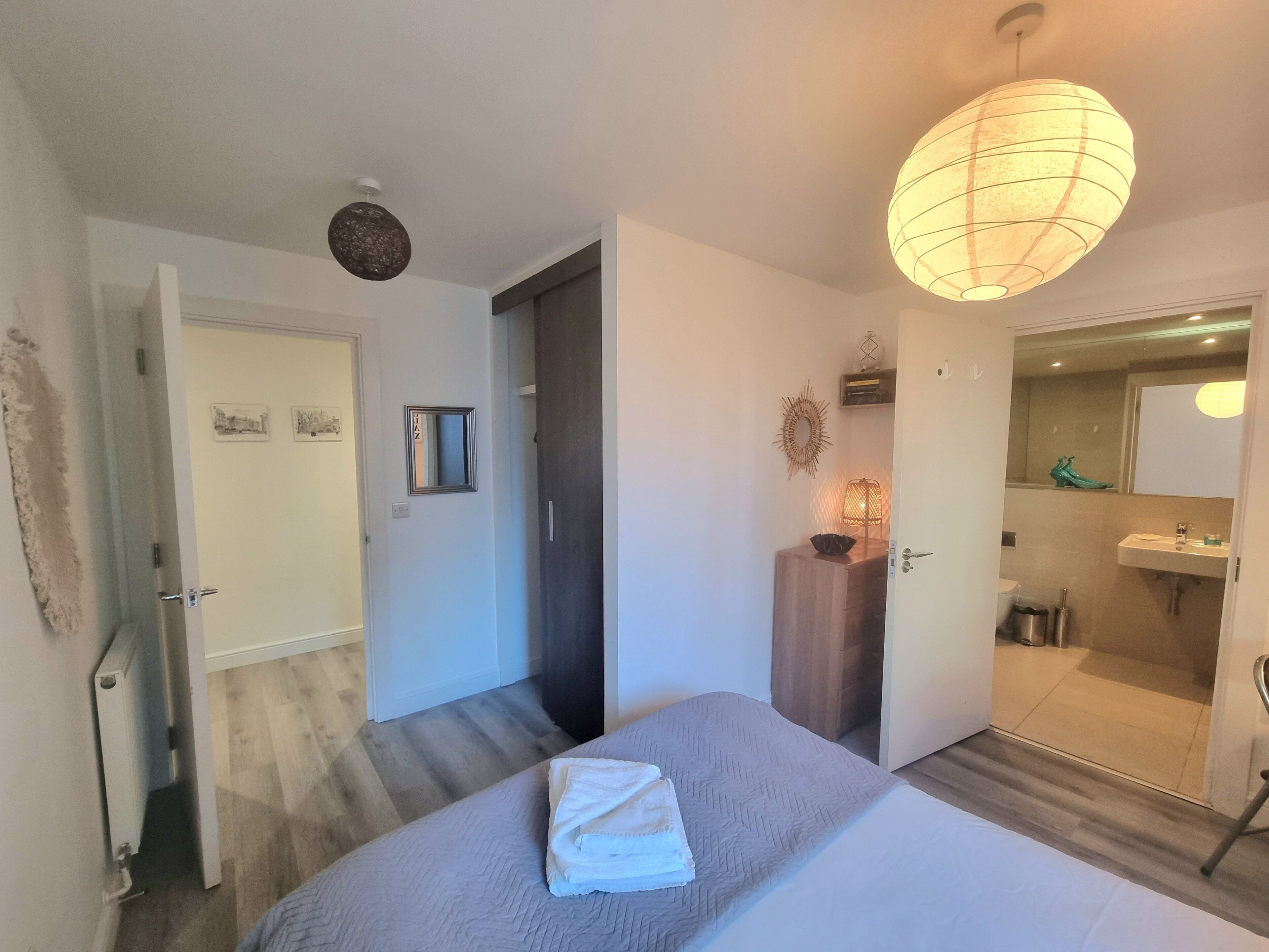 A hotel room with a bed, a bedside table, a mirror, a door leading to a bathroom, and a hanging light fixture.