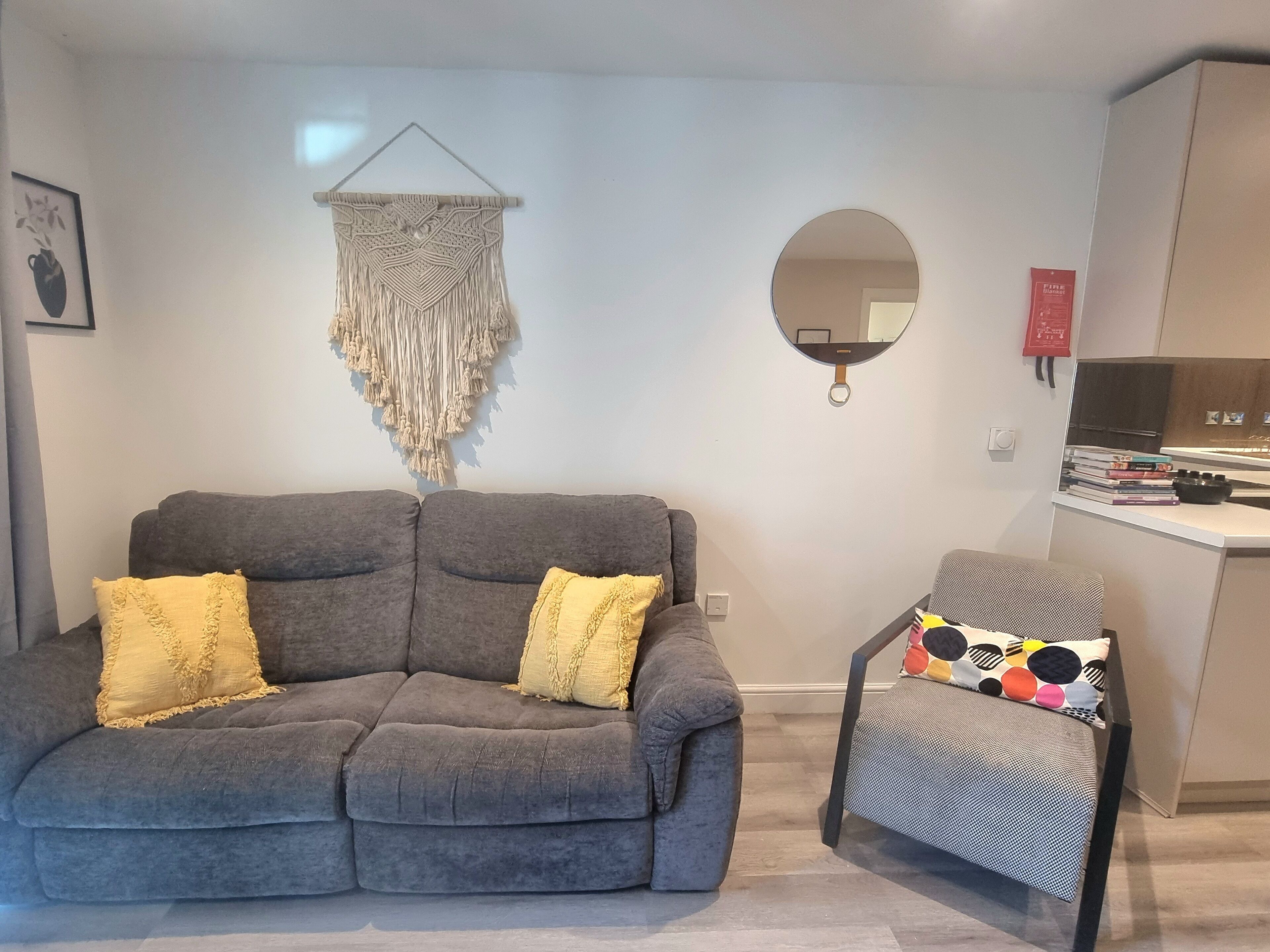 A modern living room with a grey sofa, two yellow cushions, a small round table with a colorful cushion, a macrame wall hanging, a round mirror, and a kitchen area with a fire extinguisher.