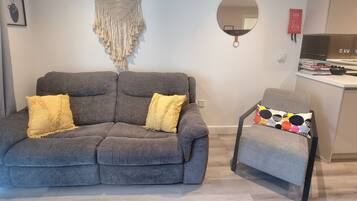 A modern living room with a grey sofa, two yellow cushions, a small round table with a colorful cushion, a macrame wall hanging, a round mirror, and a kitchen area with a fire extinguisher.