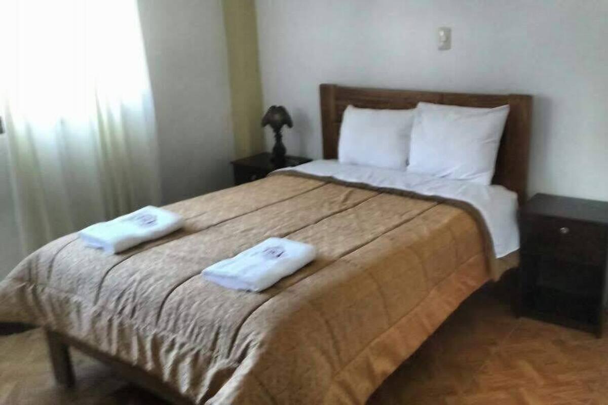 A neatly made bed with a brown bedspread and two white towels, a wooden headboard, a small bedside table with a lamp, and a dark nightstand.