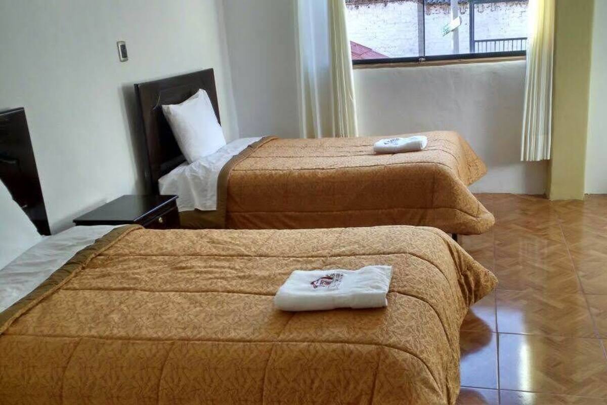 A hotel room with two beds, a wooden floor, a window with curtains, and a chair.