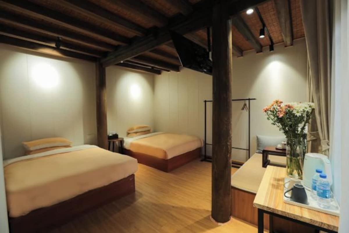 A hotel room with two beds, wooden flooring, a wooden ceiling, a small table with a vase of flowers, and a water bottle.