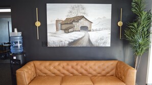 A tan leather sofa in a room with a large painting of a barn on the wall, a water dispenser, and a potted plant.