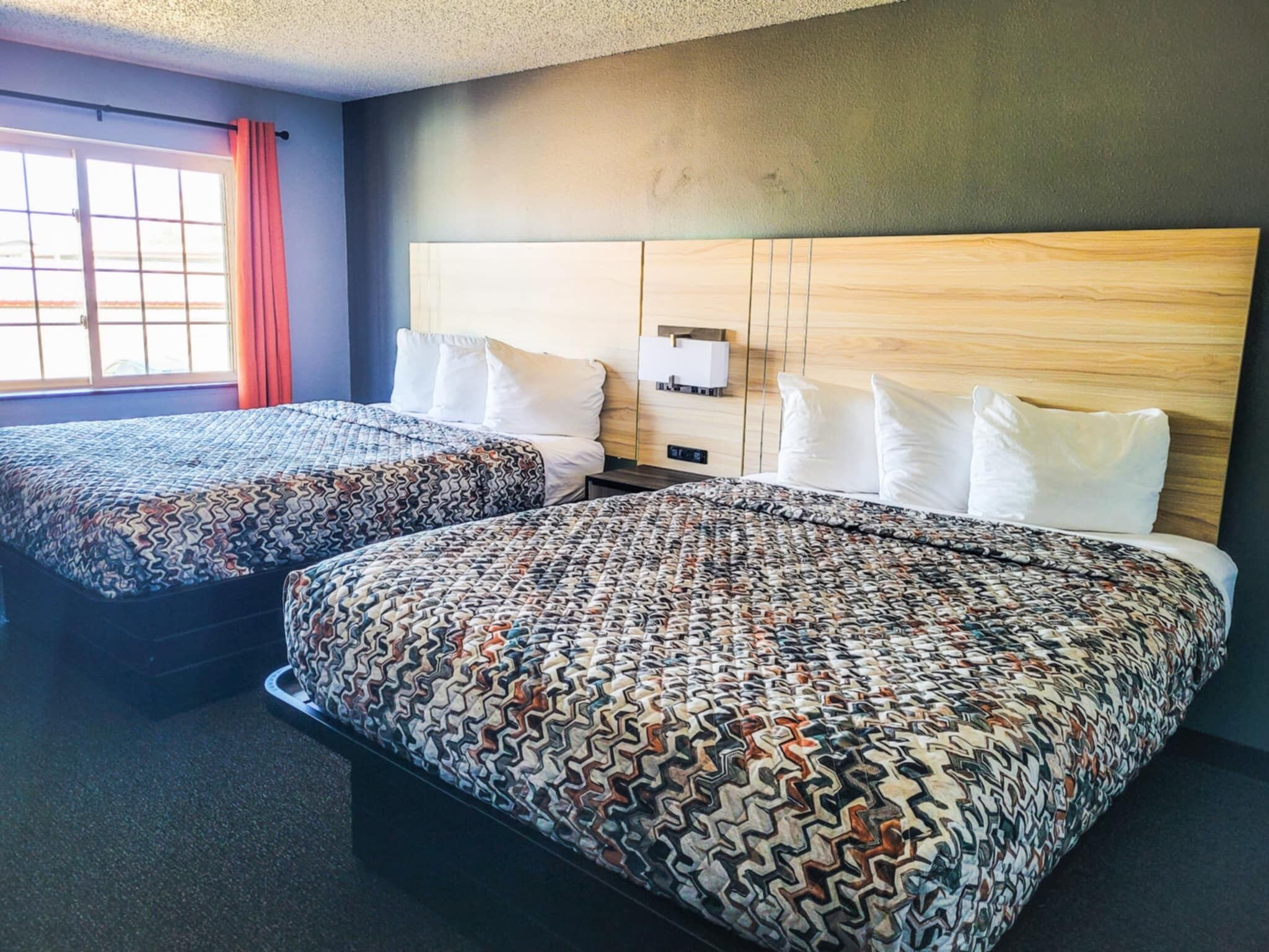 A hotel room with two beds, a wooden headboard, a window with curtains, and a small bedside table with a lamp.