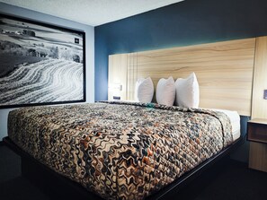 A hotel room with a large bed, a headboard with three pillows, a wall-mounted TV displaying a snowy landscape, and a wooden nightstand with a lamp. - The Northwest Inn (Spencer)
