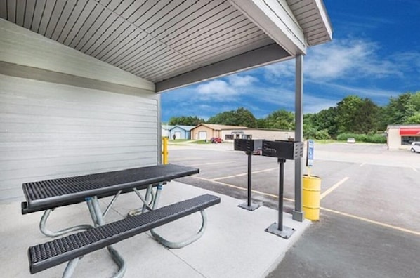 A covered picnic table with a metal frame and a bench, located in a parking lot with a building in the background. - The Northwest Inn (Spencer)