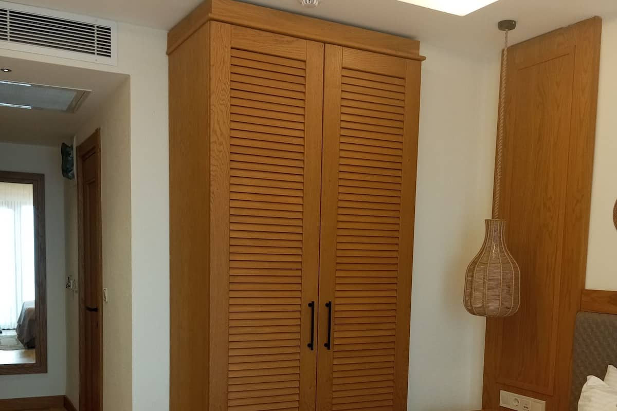Une chambre avec une grande armoire en bois, un lit recouvert d’une couvre-lit beige, une table de chevet avec une lampe et une petite table de nuit dotée d’une prise électrique.