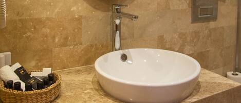 Een badkamer met een ronde spiegel, een wit wastafeltje, een handdoekhouder met opgevouwen handdoeken, een mandje met toiletartikelen en een haardroger aan de muur.
