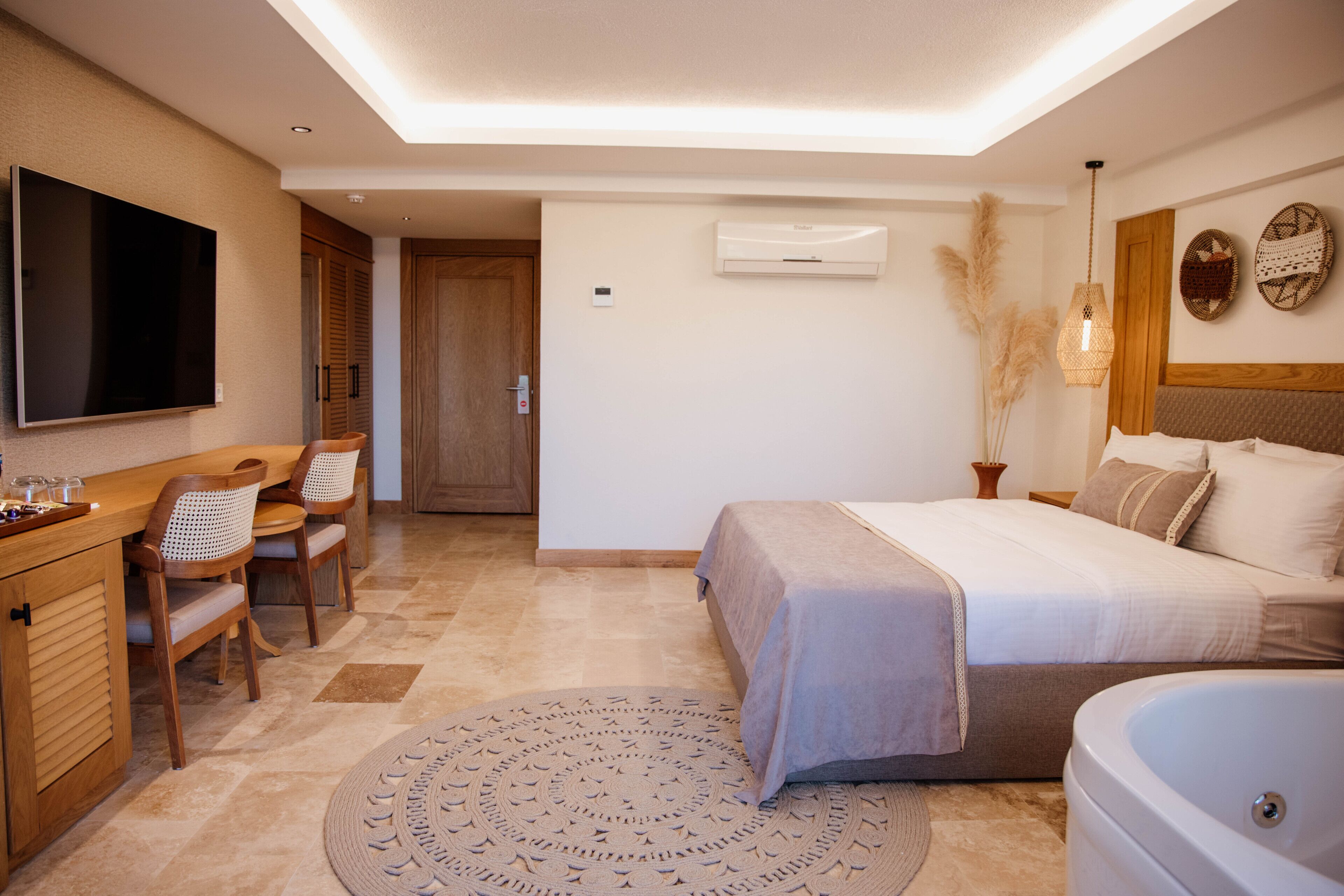 A hotel room with a bed, a desk with chairs, a TV, an air conditioning unit, and a large window.