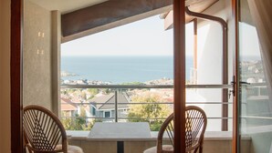 A balcony with two wicker chairs and a table, offering a view of the sea and nearby buildings.