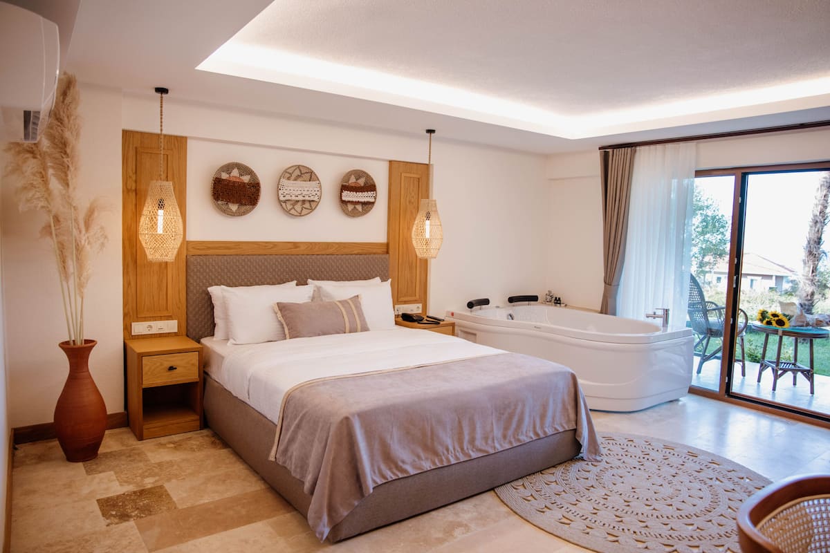 Une chambre à coucher moderne avec un grand lit, une tête de lit en bois, des tables de chevet, une petite table, une grande fenêtre avec vue et une baignoire intégrée.