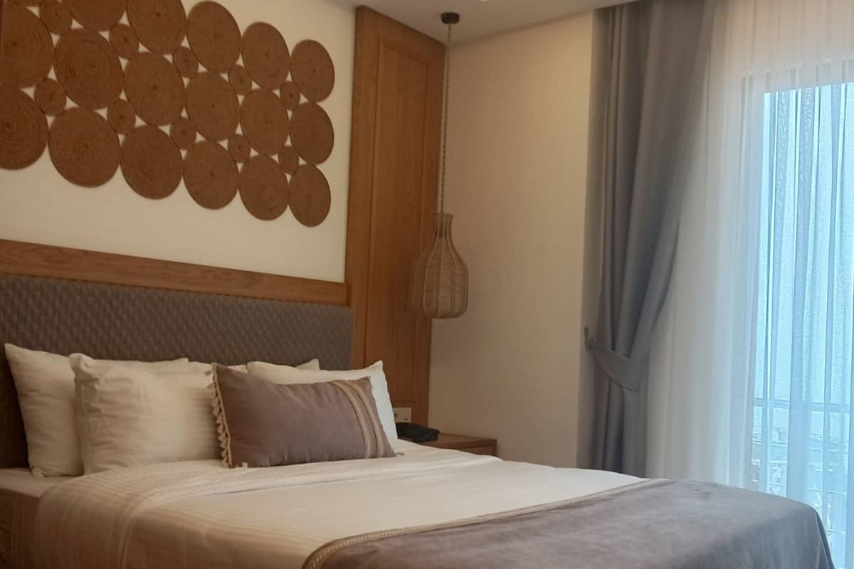 Une chambre à coucher moderne avec un grand lit, un panneau décoratif au mur, un luminaire suspendu et une vue sur l’extérieur par une fenêtre agrémentée de rideaux.
