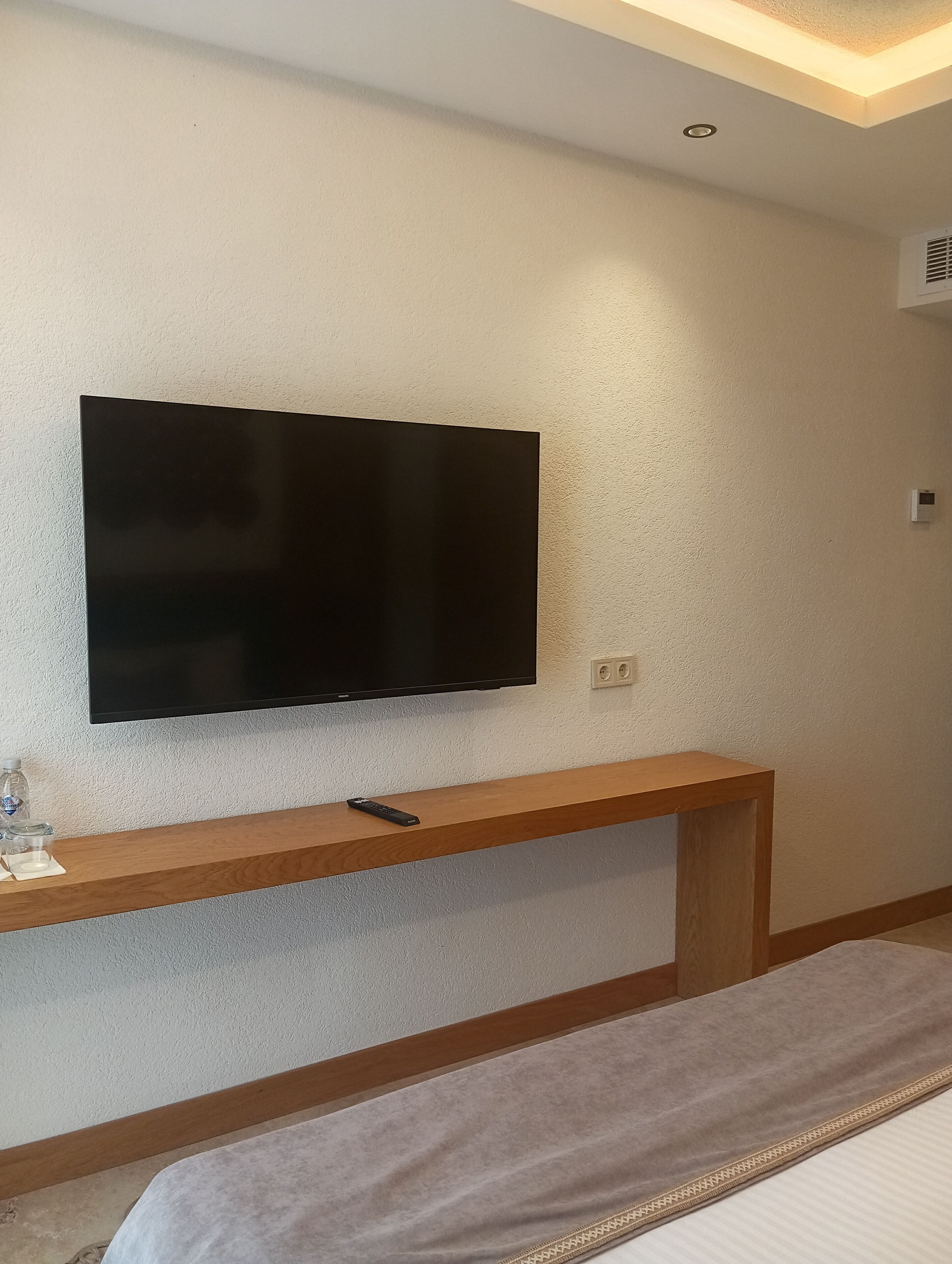 Ein modernes Zimmer mit einem an der Wand befestigten Flachbildfernseher, einer hölzernen Bank darunter und einem Bett mit gemustertem Überwurf.