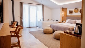 A modern hotel room with a large bed, a sitting area with a table and chairs, a bathtub, and a wooden nightstand with a kettle.