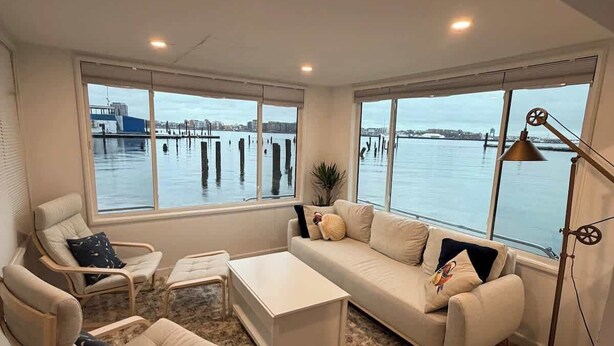 A modern living room with a large window overlooking a waterfront, a white sofa, two armchairs, a coffee table, and a floor lamp. - Endless Summer: Oceanside Views (Boston)