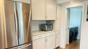 A kitchen with stainless steel appliances, white cabinetry, a microwave, and a washing machine in the adjacent room. - Endless Summer: Oceanside Views (Boston)