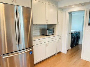 A kitchen with stainless steel appliances, white cabinetry, a microwave, and a washing machine in the adjacent room. - Endless Summer: Oceanside Views (Boston)