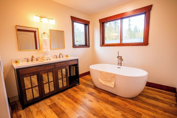 Ein Badezimmer mit einem Holzbadezimmer-Möbel, einer freistehenden Badewanne und zwei Fenstern mit Holzrahmen.