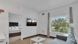 A hotel room with a bed, a desk, a chair, a small table, a sofa, a window with curtains, and a painting on the wall. - Suites at Lake Buena Vista close Disney Springs with free parking (Orlando)
