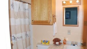 A bathroom with a shower curtain, wooden cabinets, a toilet, and a sink with toiletries. - 123 West Harbor Condo B - Beautiful Condo in the harbor with spacious deck (Port Mansfield)