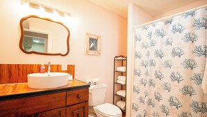 Un baño con tocador de madera, un lavabo blanco, inodoro, espejo enmarco y cortina de ducha con estampado de árboles.
