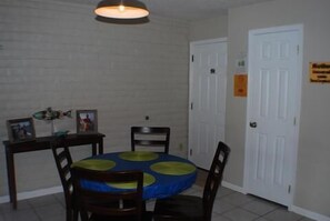 A dining area with a round table covered by a blue tablecloth with yellow circles, surrounded by wooden chairs. There is a white door with a sign on it, and a white brick wall in the background. - Fabulous Fisherman's Condo! (Port Mansfield)
