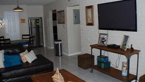 A living room with a black leather sofa, a coffee table, and a TV on a stand. There is a kitchen in the background with a refrigerator and a microwave. - Fabulous Fisherman's Condo! (Port Mansfield)