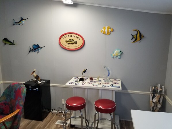Una habitación con una pared decorada con varios peces y un letrero que dice "Fish Tales Inn". Hay una mesa con fotografías y un taburete rojo.