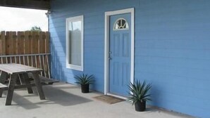 Un patio couvert avec une table et des chaises, une terrasse en bois et une maison bleue avec une porte et une fenêtre blanches.