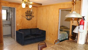 A compact living space with a dark blue sofa, a dining table with chairs, a kitchen area with cabinets, and a door leading to a bedroom with a bed. - 601 Baffin - Cozy little cottage! Screened in front porch. (Port Mansfield)