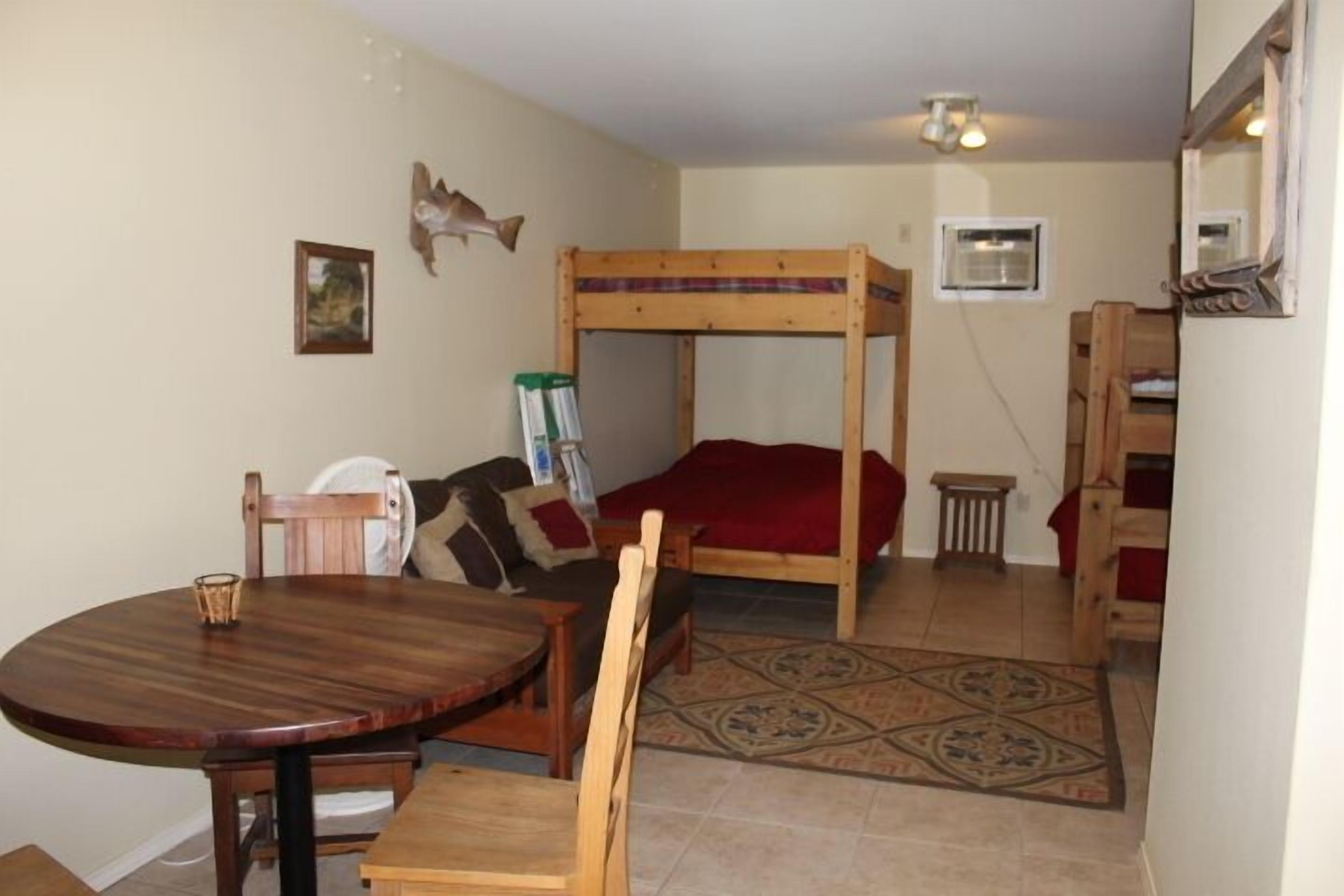 A small, cozy room with a bunk bed, a round wooden table, and a chair. There is a small rug on the floor and a framed picture on the wall.