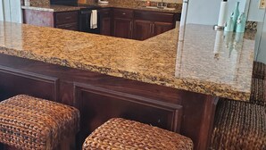Eine Küche mit dunklen Holzschränken, Granit-Arbeitsplatten und einem weißen Kühlschrank. Zwei gewebte Hocker und eine Pendelleuchte sind vorhanden.