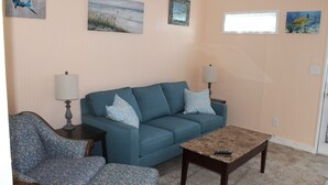 Un salon avec un canapé bleu, un fauteuil à motifs et une table basse. On y trouve des lampes fixées au mur, des œuvres d’art encadrées et une fenêtre.
