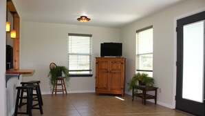 A spacious room with a tiled floor, a wooden cabinet, a television on top, and two windows with blinds. - 201 Matagorda - Cozy, Spacious Newly Remodeled Getaway! (Port Mansfield)