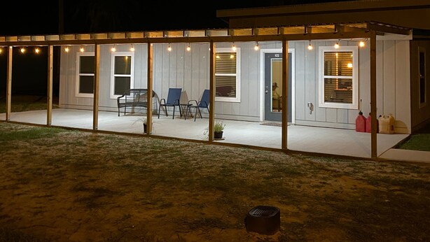 A covered outdoor patio area with string lights, a seating arrangement, and a door leading to a building. - 201 Matagorda - Cozy, Spacious Newly Remodeled Getaway! (Port Mansfield)