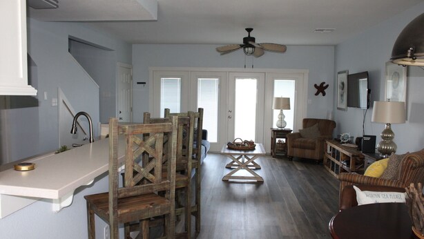 A spacious living room with a high ceiling, a ceiling fan, a dining area with wooden chairs, a sofa, and a coffee table. - Pelican Point #10 - Beautiful Complex, Gorgeous Sunrises & Easy Access (Port Mansfield)