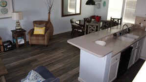A modern kitchen with a central island, a dining area with a wooden table and chairs, and a living area with a striped armchair and a lamp. - Pelican Point #10 - Beautiful Complex, Gorgeous Sunrises & Easy Access (Port Mansfield)