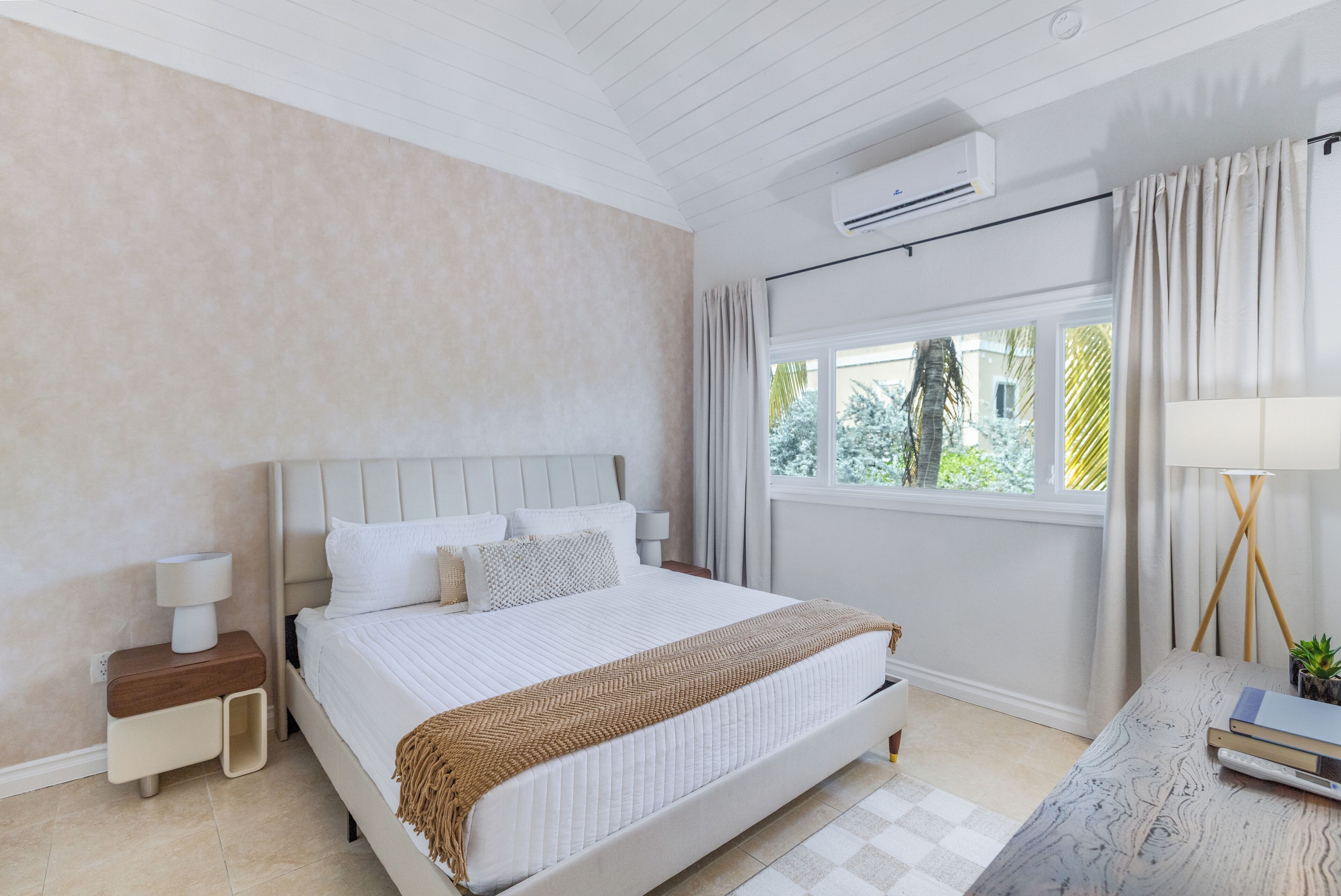 Un dormitorio con cama, mesitas de noche, equipo de aire acondicionado, ventana con cortinas y vista a zonas verdes.