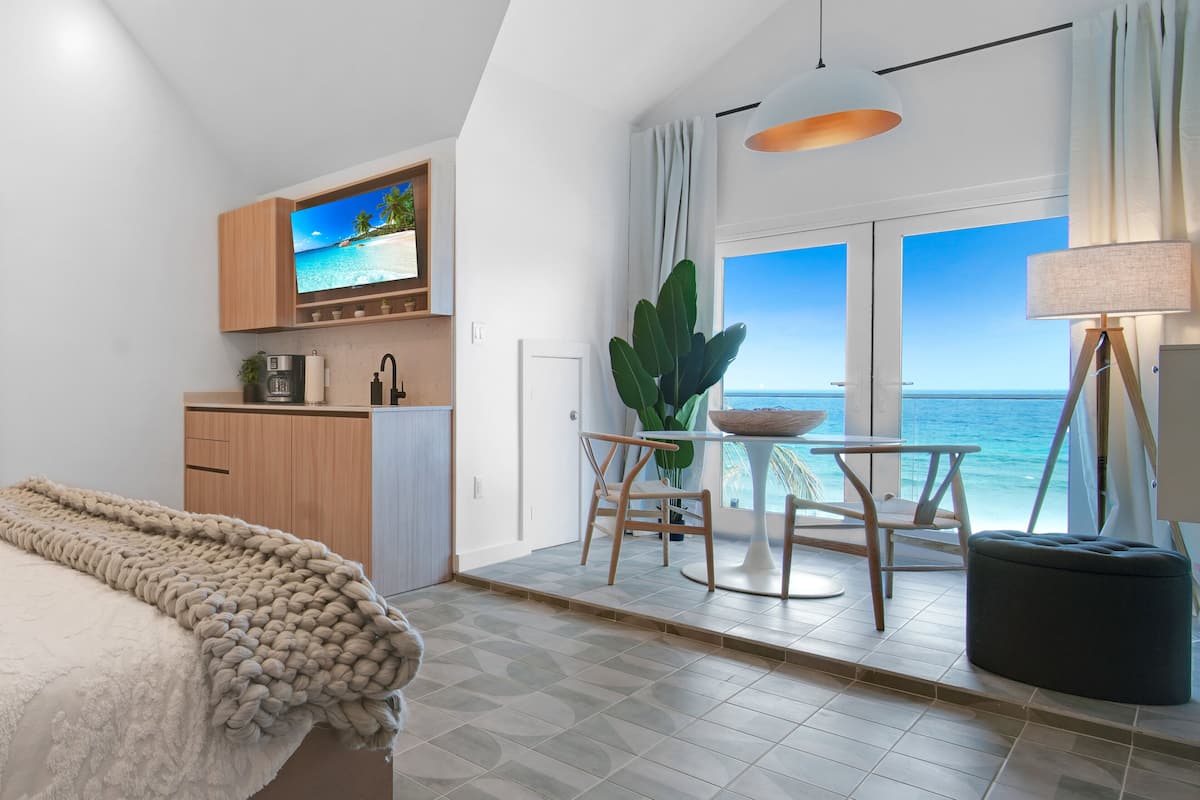 A modern room with a bed, a TV mounted on the wall, a dining area with a table and chairs, and a large window with a view of the ocean.