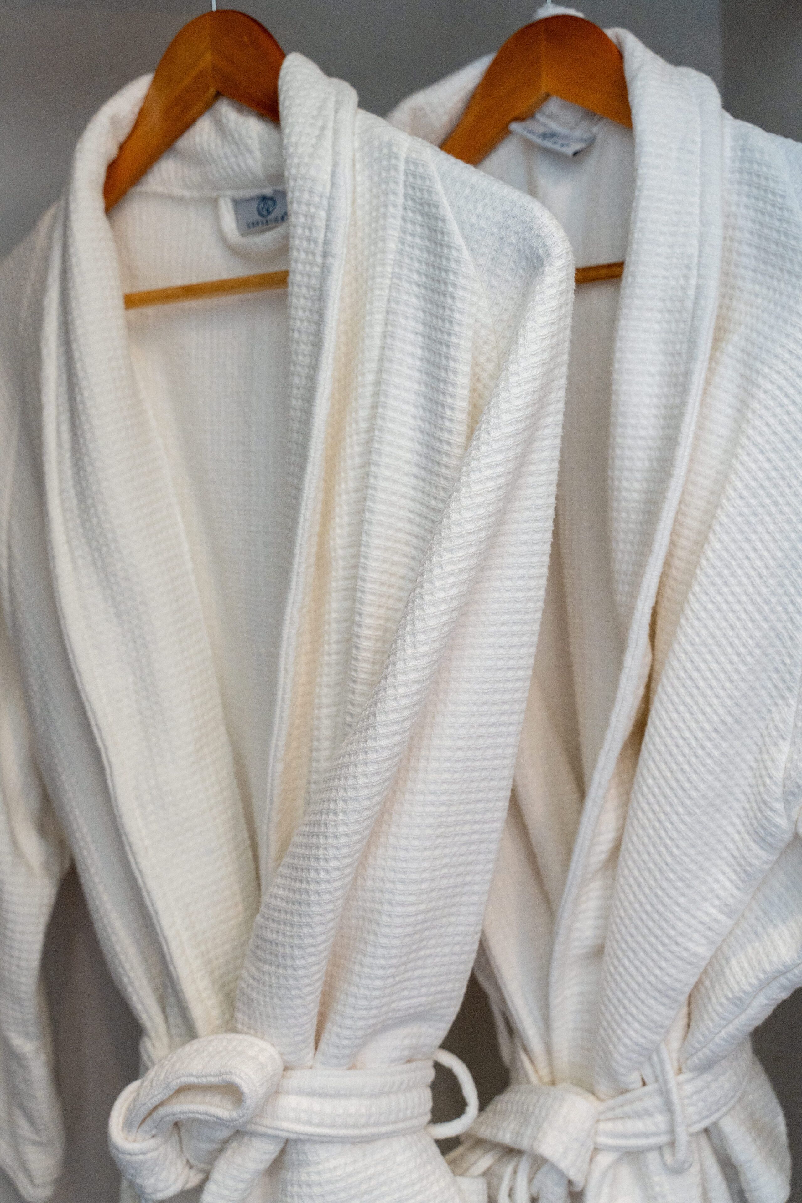 Two white bathrobes hanging on wooden hangers.