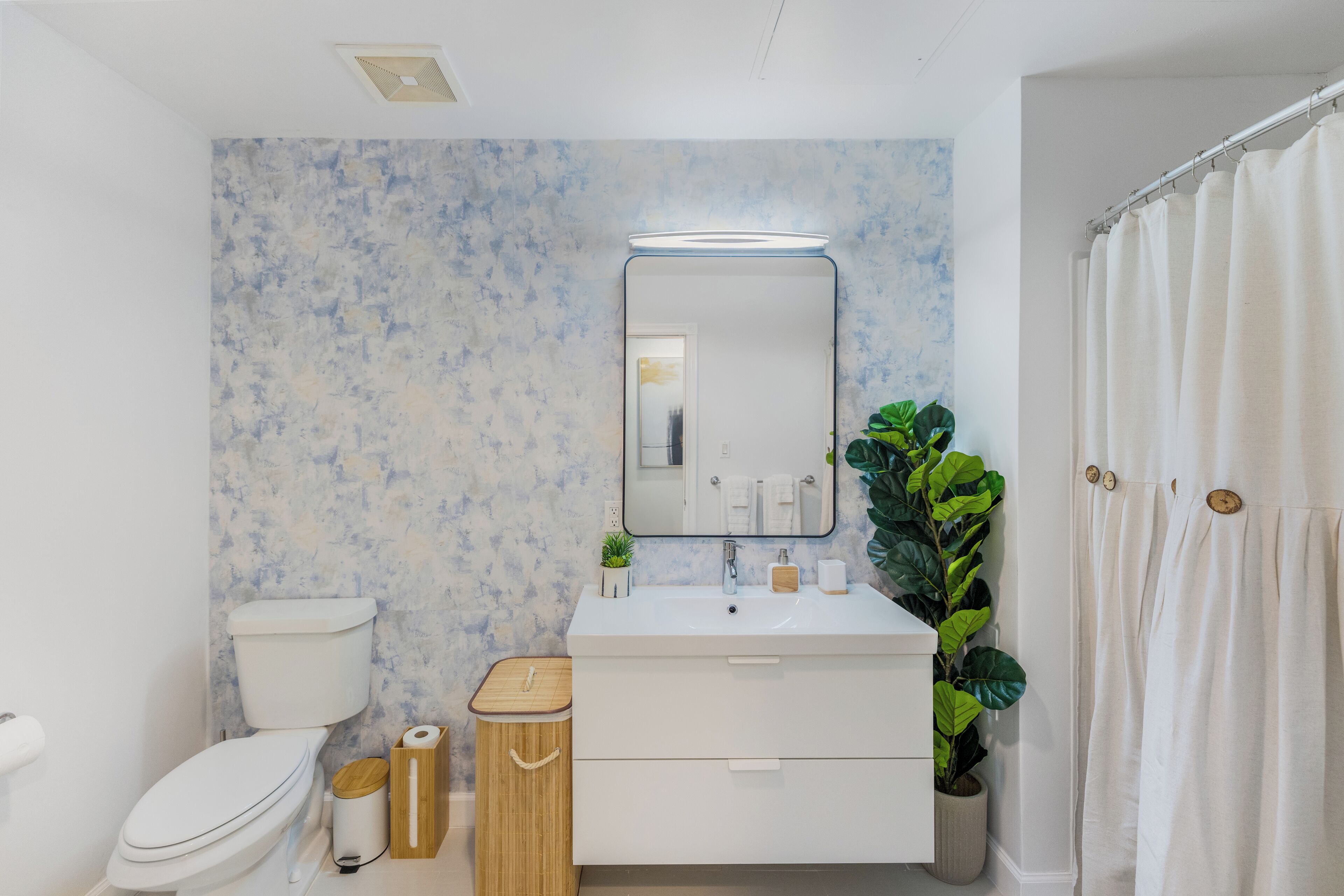 Un bagno moderno con lavandino, specchio e servizi igienici bianchi. Una pianta a destra e un cestino in legno accanto al WC.