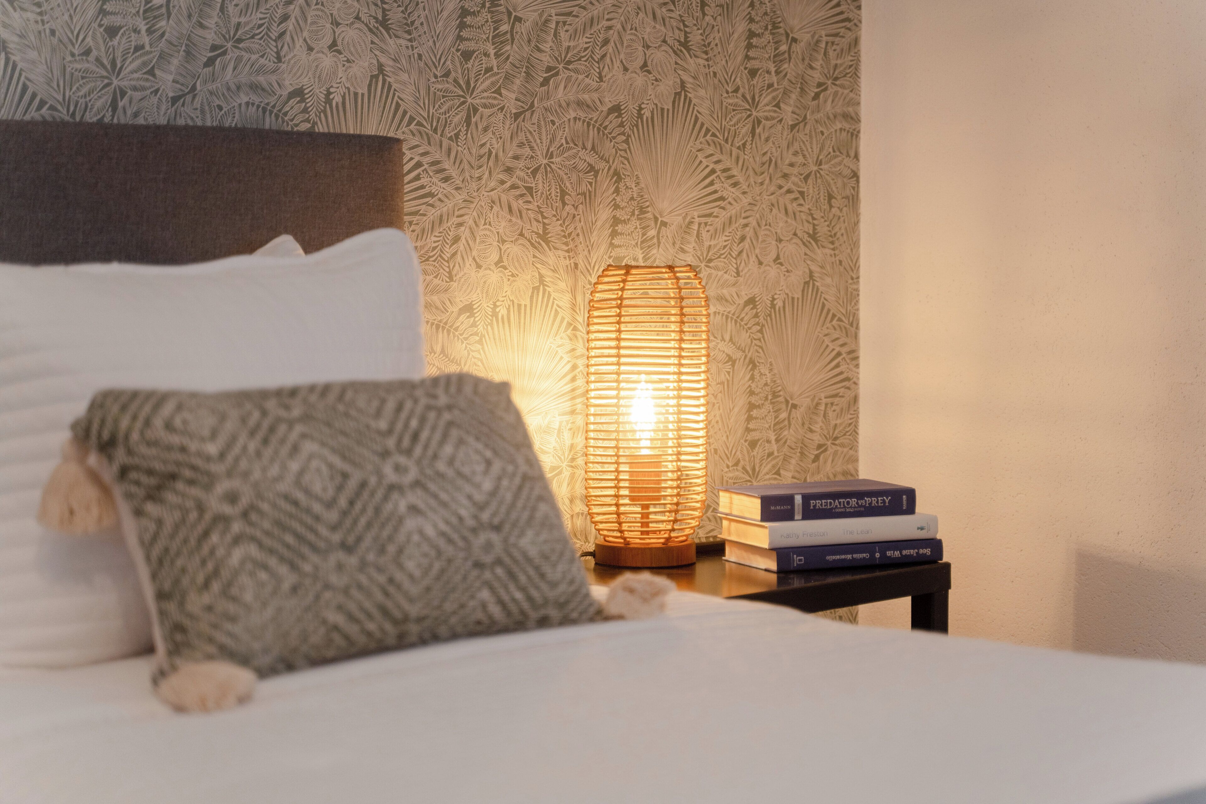 Ein ordentlich bezogenes Bett mit einem gemusterten Kissen, ein Nachttisch mit Lampe und Büchern und eine Wand mit floralem Muster.