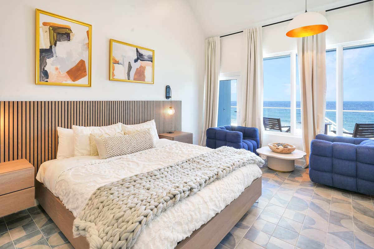 A modern hotel room with a large bed, a view of the sea, and two framed abstract paintings on the wall.